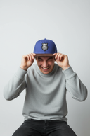 Ein Mann lacht ausgelassen, während er vor einem hellgrauen Studiohintergrund sitzt und sich mit beiden Händen eine blau-braune Kappe ins Gesicht zieht. Auf der Kappe ist prominent das gestickte Wappen eines Wildschweinkopfes zu sehen.