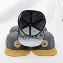 Ein Produktfoto von mehreren "Bayrischer Woid"-Kappen, das verschiedene Details zeigt. Während die unteren Kappen die Vorderseite präsentieren, ist eine Kappe in der Mitte umgedreht, um die bedruckte Unterseite des Schirms mit einem Winterwald-Foto und das Innere mit dem Snapback-Verschluss zu zeigen.