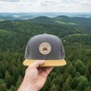 Aus der Ich-Perspektive hält eine Hand eine grau-gelbe Kappe mit einem runden "Bayrischer Woid"-Aufnäher in den Vordergrund. Im Hintergrund erstreckt sich eine weite, malerische Landschaft aus dichten Wäldern und sanften Hügeln unter einem leicht bewölkten Himmel.