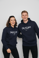 Ein Paar, ein Mann und eine Frau, lachen herzlich und ausgelassen in die Kamera. Beide tragen identische dunkelblaue Kapuzenpullover mit der weißen Aufschrift "DAHOAM" vor einem neutralen, hellgrauen Hintergrund.