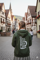 Eine Frau mit zwei blonden, geflochtenen Zöpfen steht von hinten in einer malerischen Gasse mit Kopfsteinpflaster. Sie trägt einen olivgrünen Kapuzenpullover mit einem großen "Goassmass"-Aufdruck auf dem Rücken und blickt auf die historischen Fachwerkhäuser vor ihr.