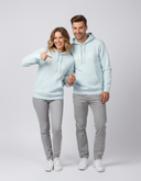 Ein Paar, ein Mann und eine Frau, posieren in passenden hellblauen Kapuzenpullovern und grauen Hosen vor einem hellgrauen Studiohintergrund. Beide lächeln in die Kamera und halten Händchen, während die Frau mit dem Finger auf den "BERG"-Schriftzug auf ihrem Pullover zeigt.