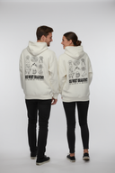 Ein Paar, ein Mann und eine Frau, steht von hinten fotografiert vor einem neutralen, hellgrauen Studiohintergrund. Sie tragen identische, cremefarbene Kapuzenpullover mit einem großen Aufdruck bayerischer Symbole auf dem Rücken, halten Händchen und blicken sich lächelnd an.