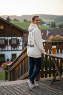 Eine lächelnde Frau mit geflochtenem Haar blickt über die Schulter, während sie auf einer Steinterrasse steht. Sie trägt einen cremefarbenen Kapuzenpullover mit einer großen Grafik auf dem Rücken. Neben ihr steht ein voller Maßkrug auf einem Holztisch, und im Hintergrund ist eine malerische Dorflandschaft mit Fachwerkhäusern zu sehen.
