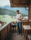 Ein Mann in einem hellen Kapuzenpullover und Jeans steht auf einem Holzbalkon und grillt auf einem kleinen Kugelgrill, während er eine Tasse in der Hand hält. Im Hintergrund erstreckt sich eine malerische alpine Tallandschaft mit Bergen.