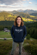 Eine Frau lacht herzlich in die Kamera, während sie auf einer Anhöhe vor einer atemberaubenden Bergkulisse steht. Sie trägt einen dunkelblauen Kapuzenpullover mit der Aufschrift "DAHOAM" und blickt über ein weites Tal mit Wäldern und einem kleinen Dorf in der Ferne.