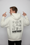 Ein Mann, von hinten fotografiert, präsentiert den großen Aufdruck auf seinem cremefarbenen Kapuzenpullover. Er blickt mit einem breiten Lächeln über die Schulter in die Kamera und zeigt spielerisch mit beiden Zeigefingern nach oben.