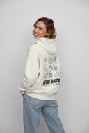 Eine lächelnde Frau blickt über ihre Schulter in die Kamera und präsentiert dabei die Rückseite ihres cremefarbenen Kapuzenpullovers mit einem großen Aufdruck bayerischer Symbole. Sie trägt weite Jeans und hat die Hände in den Hosentaschen.