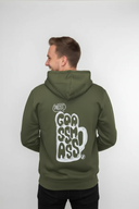 Ein Mann, von der Seite von hinten fotografiert, präsentiert den großen weißen "Goassmass"-Aufdruck auf seinem olivgrünen Kapuzenpullover. Er blickt mit einem freundlichen Lächeln über die Schulter.