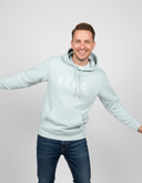 Ein Mann mit einem breiten, fröhlichen Lächeln posiert in einem hellblauen Kapuzenpullover. Er hat die Arme zur Seite ausgestreckt und blickt direkt in die Kamera.