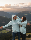 Ein Paar in passenden hellblauen Kapuzenpullovern hat auf einem Berggipfel bei Sonnenuntergang sichtlich Spaß. Die Frau zieht ihrem lachenden Partner spielerisch die Kapuze über den Kopf, während im Hintergrund eine atemberaubende Bergkulisse zu sehen ist.