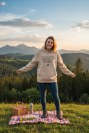 Eine glückliche Frau in einem beigen Kapuzenpullover steht mit offenen Armen auf einer Bergwiese bei Sonnenuntergang. Vor ihr auf einer karierten Decke ist ein Picknick mit Korb und Thermoskanne ausgebreitet, während im Hintergrund eine malerische Landschaft aus Hügeln und Bergen liegt.