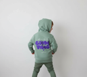 Ein kleiner Junge, von hinten fotografiert, präsentiert den großen lila "GUADE VIBES"-Aufdruck auf seinem salbeigrünen Kapuzenpullover. Er hat die Kapuze aufgesetzt und blickt über die Schulter.