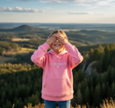 Ein lächelndes Mädchen steht auf einem Aussichtspunkt und hält sich mit beiden Händen die Augen zu. Es trägt einen pinken "LAUSDEANDL"-Kapuzenpullover vor einer malerischen Kulisse aus bewaldeten, sanften Hügeln bei Sonnenuntergang.
