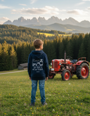Ein kleiner Junge, von hinten gesehen, steht in einem dunkelblauen Kapuzenpullover auf einer blühenden Almwiese. Er blickt auf eine beeindruckende Bergkette in der Ferne, während neben ihm ein alter roter Traktor parkt, passend zum Motiv auf seinem Rücken.