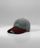 Ein professionelles Produktfoto einer zweifarbigen Baseball-Kappe. Die Kappe hat eine graue Krone und einen weinroten Schirm. Auf der Vorderseite ist ein buntes Logo aufgestickt, das stilisierte Berge und ein Gewässer zeigt. Die Kappe steht auf einer spiegelnden Oberfläche vor einem hellen Studiohintergrund, was eine deutliche Reflexion erzeugt.