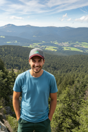 Ein lächelnder junger Mann steht auf einem Aussichtspunkt in den Bergen. Er trägt ein blaues T-Shirt und eine graue Kappe mit weinrotem Schirm. Im Hintergrund erstreckt sich eine weite Landschaft aus bewaldeten Hügeln und Bergen unter einem leicht bewölkten Himmel.
