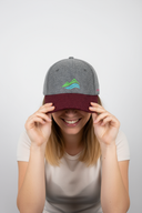 Eine lächelnde Frau sitzt vor einem hellgrauen Studiohintergrund und zieht sich mit beiden Händen eine graue Kappe mit weinrotem Schirm und einem bunten Berg-Logo über die Augen. Ihr Lächeln ist unter dem Schirm sichtbar.