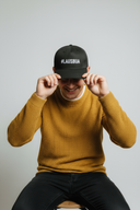 Eine lächelnde Frau sitzt auf einem Hocker vor einem hellgrauen Studiohintergrund. Sie setzt sich gerade eine schwarze Kappe mit dem weißen Schriftzug "#LAUSDEANDL" auf und blickt dabei freundlich in die Kamera.