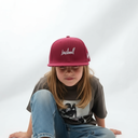 Ein Mädchen mit langen Haaren sitzt vor einem hellgrauen Studiohintergrund und blickt nach unten. Sie trägt eine weinrote Kappe mit dem weißen, kursiven Schriftzug "Lausdeandl".