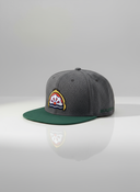 Ein professionelles Produktfoto einer grau-grünen Kappe mit flachem Schirm. Auf der Vorderseite befindet sich ein aufgesticktes, farbenfrohes Brauerei-Logo. Die Kappe steht auf einer spiegelnden Oberfläche vor einem hellen Studiohintergrund, was eine deutliche Reflexion erzeugt.