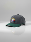 Ein professionelles Produktfoto einer grau-grünen Kappe mit gebogenem Schirm. Auf der Vorderseite befindet sich ein aufgesticktes, farbenfrohes Brauerei-Logo. Die Kappe steht auf einer spiegelnden Oberfläche vor einem hellen Studiohintergrund, was eine deutliche Reflexion erzeugt.