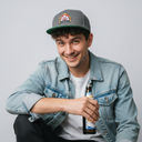 Ein junger Mann in einer Jeansjacke sitzt vor einem hellgrauen Studiohintergrund und lächelt freundlich in die Kamera. Er trägt eine grau-grüne Kappe mit einem Brauerei-Logo und hält eine Flasche Bier in der Hand.