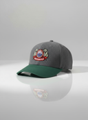 Ein professionelles Produktfoto einer grau-grünen Kappe mit gebogenem Schirm. Auf der Vorderseite befindet sich ein aufgesticktes, farbenfrohes "Mühlbauer"-Logo mit zwei Bären. Die Kappe steht auf einer spiegelnden Oberfläche vor einem hellen Studiohintergrund, was eine deutliche Reflexion erzeugt.