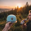 Aus der Ich-Perspektive hält eine Hand eine hellblaue "OIMARA"-Kappe in den Vordergrund. Im Hintergrund trinkt ein Mann aus einer Bierflasche, während die Sonne hinter einer malerischen Landschaft aus Wäldern und Bergen untergeht.
