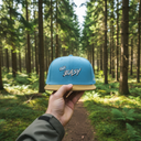 Aus der Ich-Perspektive hält eine Hand eine hellblaue Kappe mit dem "Ois Easy"-Schriftzug in den Vordergrund. Im Hintergrund erstreckt sich ein sonnendurchfluteter Waldweg, der von hohen Nadelbäumen und grünem Farn gesäumt ist.