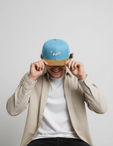 Ein Mann mit einem breiten Lächeln sitzt vor einem hellgrauen Studiohintergrund und zieht sich mit beiden Händen eine hellblaue Kappe mit braunem Schirm und dem Schriftzug "Ois Easy" ins Gesicht.