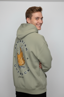 Ein junger Mann, von der Seite von hinten fotografiert, präsentiert den großen "Bavarian Outback"-Aufdruck mit Lagerfeuer-Motiv auf seinem salbeigrünen Kapuzenpullover. Er blickt mit einem breiten Lächeln über die Schulter.