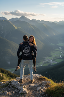 Auf einem Berggipfel mit atemberaubender Aussicht auf ein Tal und eine Bergkette trägt ein Mann eine Frau huckepack. Die Frau lacht ausgelassen, während der Mann den "WANDERLUST"-Kapuzenpullover trägt, dessen Aufdruck zur Kamera zeigt.