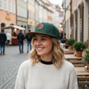 Eine lächelnde junge Frau mit blonden, gewellten Haaren steht in einer Gasse in einer europäischen Altstadt. Sie trägt einen cremefarbenen Strickpullover sowie eine grüne Kappe und blickt an den Tischen eines Straßencafés vorbei in die Ferne.