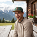 Ein herzlich lachender junger Mann sitzt an einem Holztisch auf der Terrasse einer rustikalen Almhütte. Er trägt einen beigen Strickpullover und eine grüne Kappe und blickt zur Kamera. Im Hintergrund erstreckt sich eine beeindruckende, schneebedeckte Bergkette.