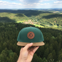 Aus der Ich-Perspektive hält eine Hand eine grüne Kappe mit einem runden Lederaufnäher in den Vordergrund. Im Hintergrund erstreckt sich eine weite, malerische Landschaft aus dichten Wäldern und einem kleinen Dorf in einem Flusstal.