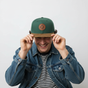 Ein Mann mit einem breiten Lächeln sitzt vor einem hellgrauen Studiohintergrund und zieht sich mit beiden Händen eine grüne Kappe mit braunem Wildlederschirm und einem runden Lederaufnäher ins Gesicht.