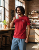 Ein lächelnder Mann mit dunklen, lockigen Haaren steht in einem sonnendurchfluteten Café mit Backsteinwänden. Er trägt ein rotes T-Shirt und hebt eine weiße Kaffeetasse zum Trinken, während er zur Seite blickt.