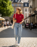 Eine lächelnde junge Frau schlendert an einem sonnigen Tag eine Kopfsteinpflasterstraße entlang. Sie trägt ein rotes T-Shirt, helle Jeans und eine Stofftasche über der Schulter, während sie einen Kaffeebecher in der Hand hält und zur Seite blickt.