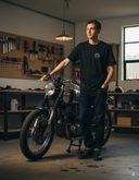 Ein junger Mann in einem schwarzen T-Shirt und dunklen Jeans steht in einer Werkstatt neben einem klassischen Motorrad. Eine Hand ruht auf dem Lenker, während er nachdenklich zur Seite blickt. Im Hintergrund sind eine Werkbank und eine Wand mit aufgehängten Werkzeugen zu sehen.