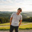 Ein fröhlich lachender Mann steht mit den Händen in den Hosentaschen auf einer Anhöhe. Er trägt eine Kappe mit der Aufschrift "Spinnradl". Im Hintergrund erstreckt sich eine weite Landschaft aus grünen Wiesen und bewaldeten Hügeln im Licht der Abendsonne.