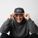 Ein Mann mit einem breiten Lächeln sitzt vor einem hellgrauen Studiohintergrund. Er setzt sich gerade eine dunkelgraue Kappe mit braunem Schirm und dem weißen Schriftzug "Spinnradl" auf und blickt dabei direkt in die Kamera.