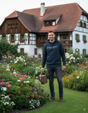 Ein lächelnder Mann in einem dunkelblauen Kapuzenpullover steht auf einem gepflegten Rasenweg inmitten eines prächtigen Blumengartens. Er hält eine Gießkanne in der Hand und blickt in die Kamera. Im Hintergrund steht ein großes, traditionelles Fachwerkhaus.