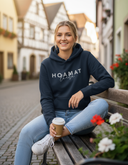 Eine lächelnde junge Frau mit blonden Haaren sitzt auf einer Holzbank in einer malerischen Gasse mit Kopfsteinpflaster. Sie trägt einen dunkelblauen Kapuzenpullover mit der Aufschrift "HOAMAT liebe" und hält einen Kaffeebecher in der Hand, während sie direkt in die Kamera blickt.