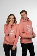 Ein Paar in passenden lachsfarbenen Kapuzenpullovern posiert lachend vor einem hellgrauen Hintergrund. Die Frau hält sich amüsiert die Hand vor den Mund, während der Mann sie mit einem verschmitzten Grinsen von der Seite ansieht.