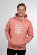 Ein junger Mann mit einem breiten, sympathischen Lächeln posiert in einem lachsfarbenen Kapuzenpullover. Er hat die Hände in der vorderen Bauchtasche des Hoodies und blickt direkt in die Kamera.