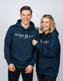 Ein junger Mann und eine Frau in passenden dunkelblauen Kapuzenpullovern mit der Aufschrift "HOAMAT liebe" lachen herzlich und ausgelassen in die Kamera.