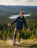 Ein junger Mann in einem dunkelblauen "HOAMAT liebe"-Kapuzenpullover steht mit ausgebreiteten Armen und einem Ausdruck freudiger Überraschung auf einer Anhöhe. Hinter ihm erstreckt sich eine weite Landschaft mit einem See, der von dichten Wäldern und Hügeln umgeben ist.