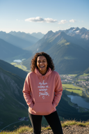 Eine Frau mit dunklen, lockigen Haaren lacht ausgelassen auf einem Berggipfel. Sie trägt einen lachsfarbenen Kapuzenpullover mit der Aufschrift "mehra schmusn, weniga grantln" und hat die Hände in der Bauchtasche. Hinter ihr eröffnet sich eine atemberaub's eyeende Aussicht auf ein tiefes Tal mit Seen und einer schneebedeckten Bergkette.