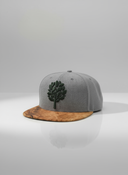 Ein professionelles Produktfoto einer grauen Kappe mit einem flachen Schirm in Holzoptik. Auf der Vorderseite befindet sich ein erhabenes, dunkelgrünes Baum-Logo. Die Kappe steht auf einer spiegelnden Oberfläche vor einem hellen Studiohintergrund, was eine deutliche Reflexion erzeugt.